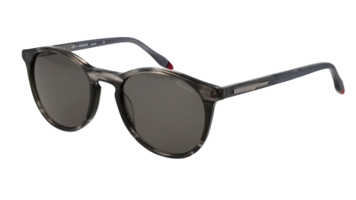 A pair of designer sunglasses with a translucent dark gray and black tortoiseshell frame and dark gray lenses, featuring a silver-tone metal logo plate on the temple arm.